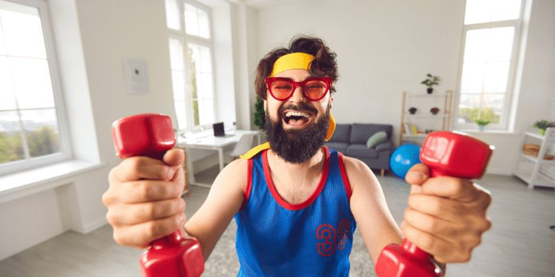 Bearded person in sunglasses exercises with red dumbbells, smiling.